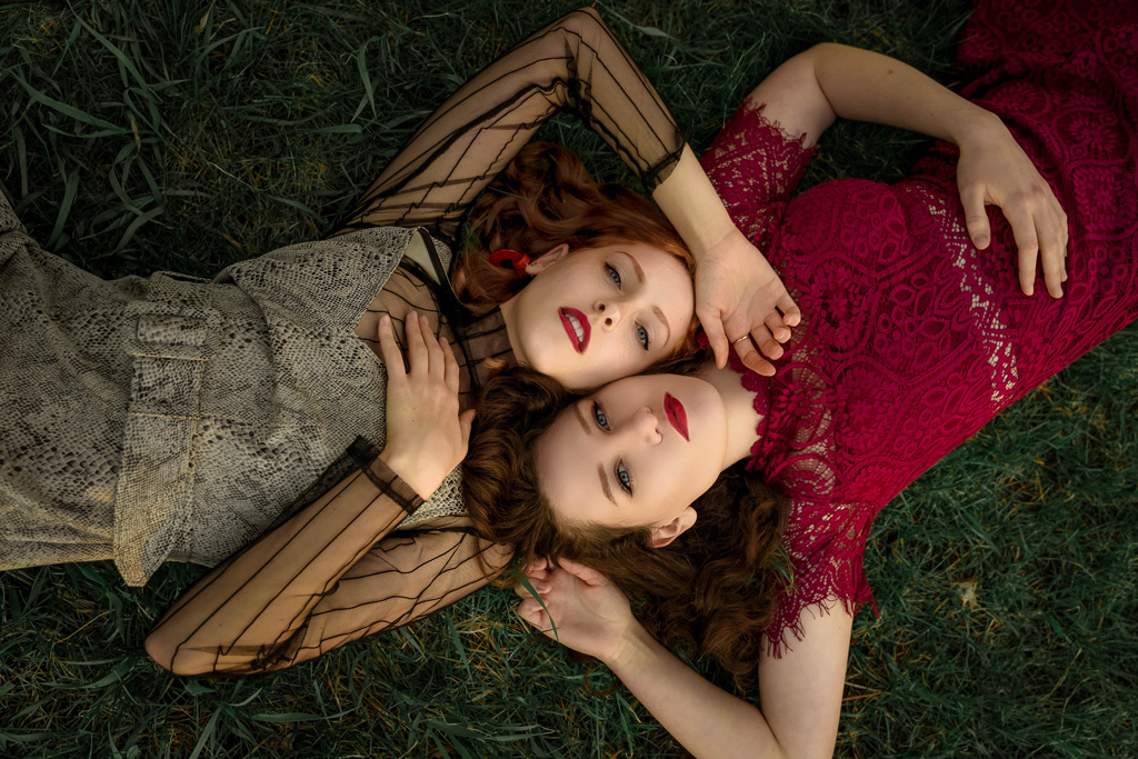 Portrait shot from above, two women laying down in the grass in opposing directions, their heads close next to each other, one wearing a red the other a grey lace dress
