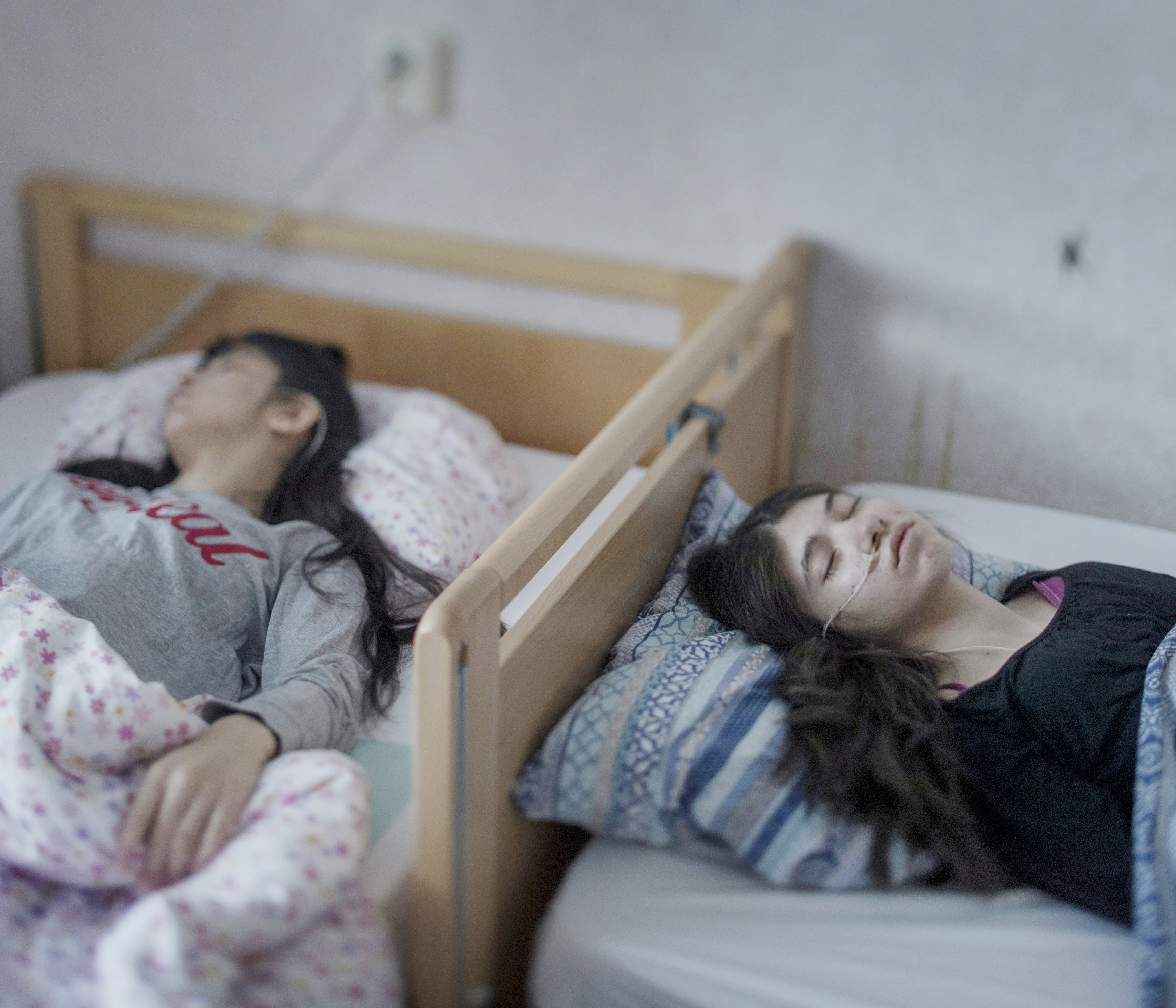 Two teenage girls lie asleep in beds touching each other, with hospital tubes in their noses.