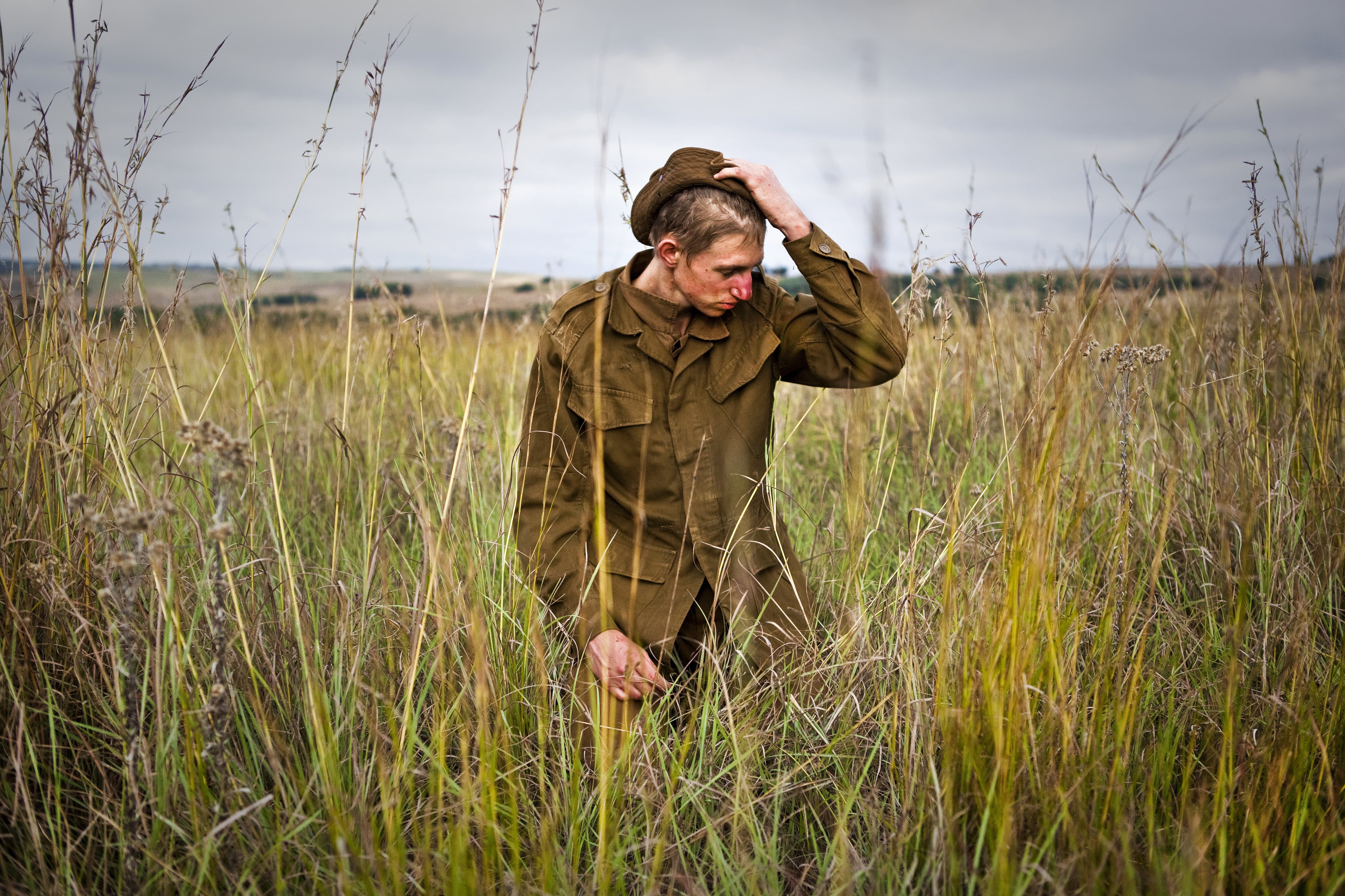 A young blonde boy in military uniform stands in a field of long grass, adjusting his hat.