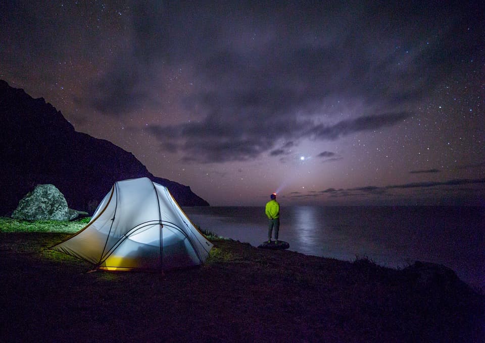 man standing in front of a well-lit tent looking at the stars - night landscape photography