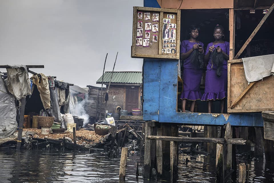 Nigeria, Lagos Two women, dressed in purple and holding weaves, stand in the doorway of a hair and beauty saloon, one of several such waterside establishments in Makoko. They are usually very busy on Sunday, when the women of the local community are preparing for church or other ceremonies. The Makoko slum is a community, under threat from eviction, built over the polluted waters of the Lagos Lagoon.