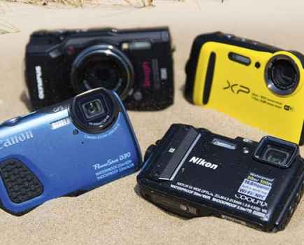 Four compact cameras in the sand: two black, one yellow and one blue