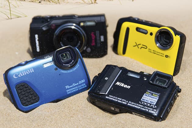 Four compact cameras in the sand: two black, one yellow and one blue