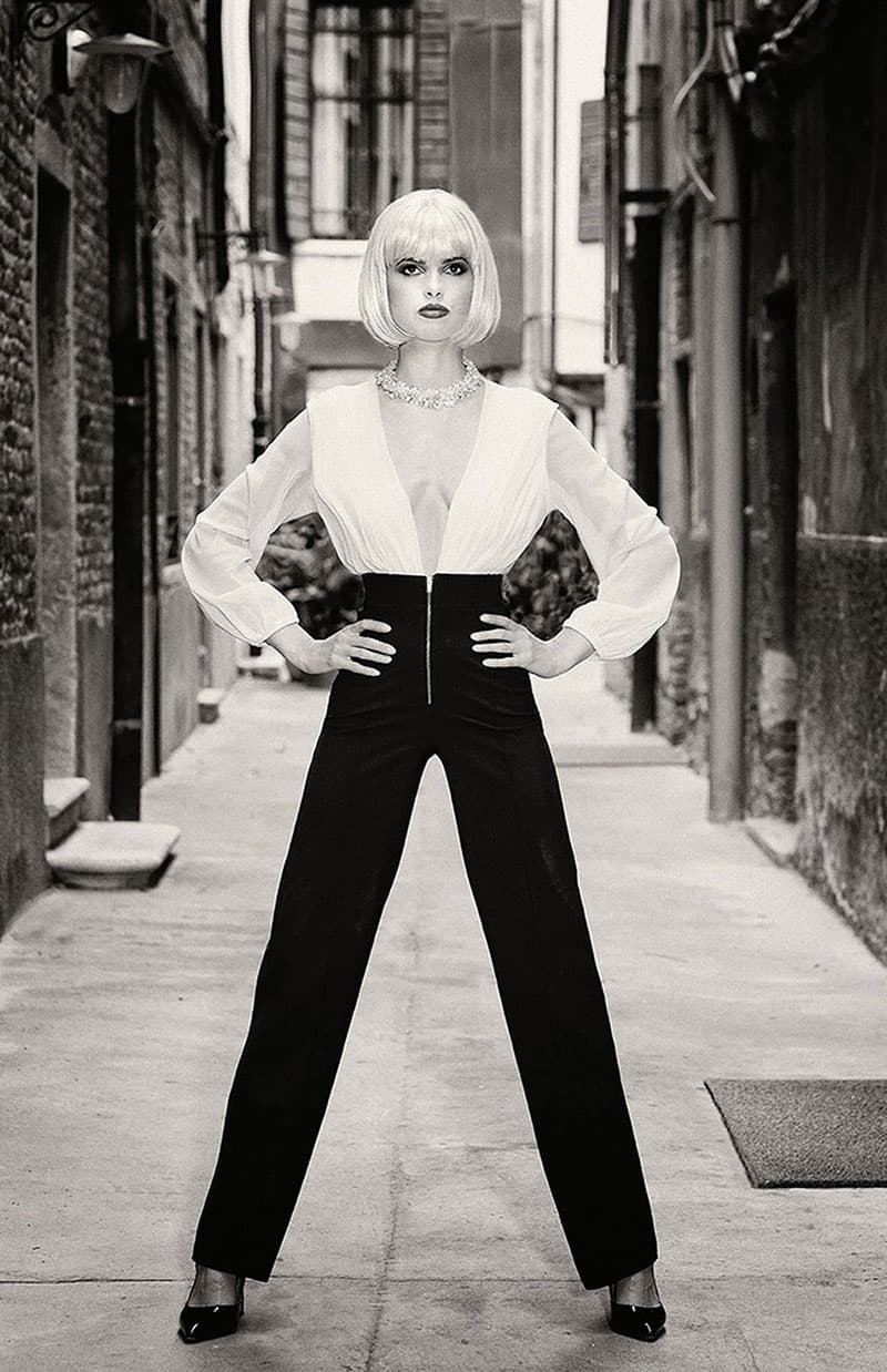 Yerbury portraits bespoke background. Model wearing black trousers and white blouse posing with her hands on her hips in an alleyway.