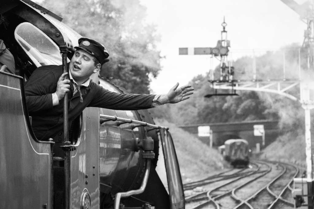 Canon EOS 90D sample image, black and white, steam train driver leans out from the train one hand stretched, in the background we can see multiple train tracks and trees along them