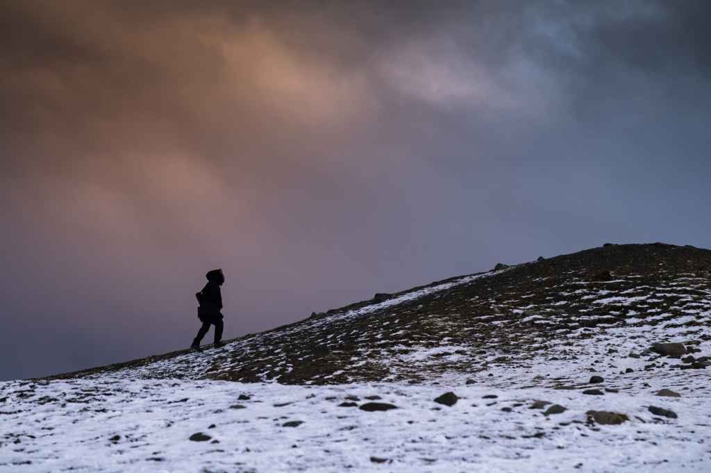 person walking up snowy hill with backdrop of orange clouds