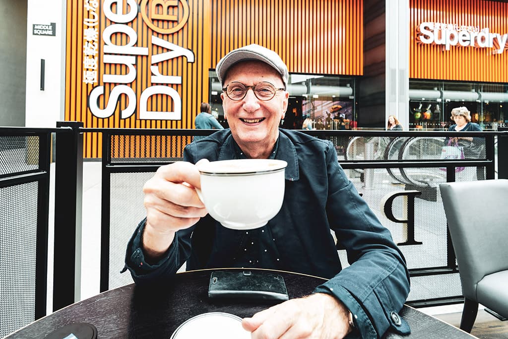 Man raising his teacup to the camera in a shopping mall with a Superdry store behind