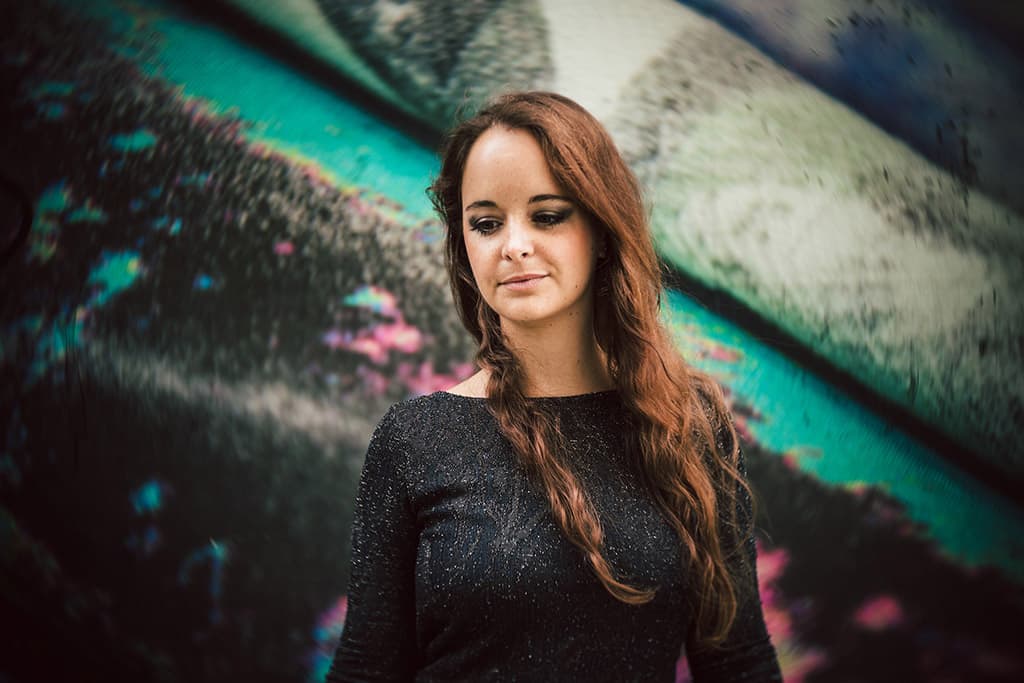 A brunette woman with dark eye makeup stood infront of a graphic coloured wall