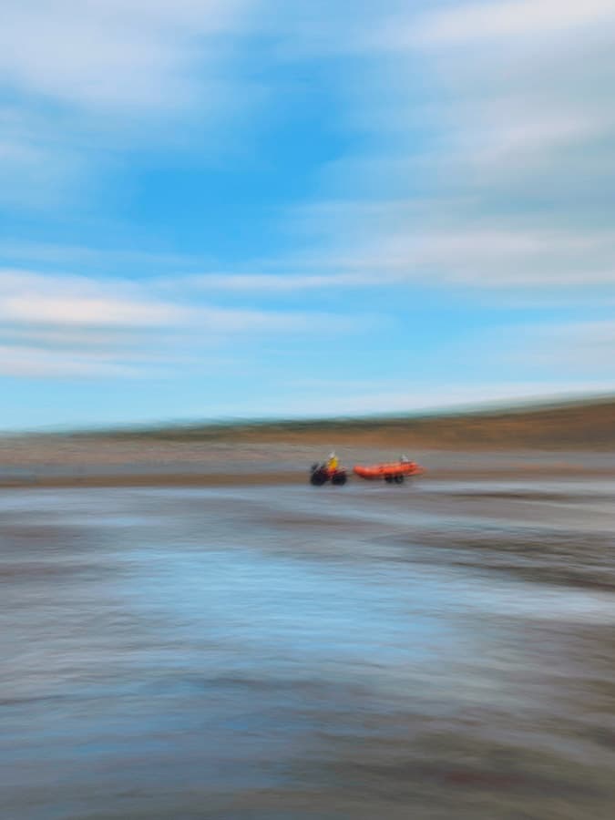 A painterly scene with motion blur, sky and land half the image, in the middle a person on a small tractor towing a red boat. Widemouth Bay, Bude, Cornwall. smartphone photos