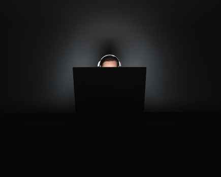 A person sat at a desk in the dark with the top of their head peeping from bright computer screen