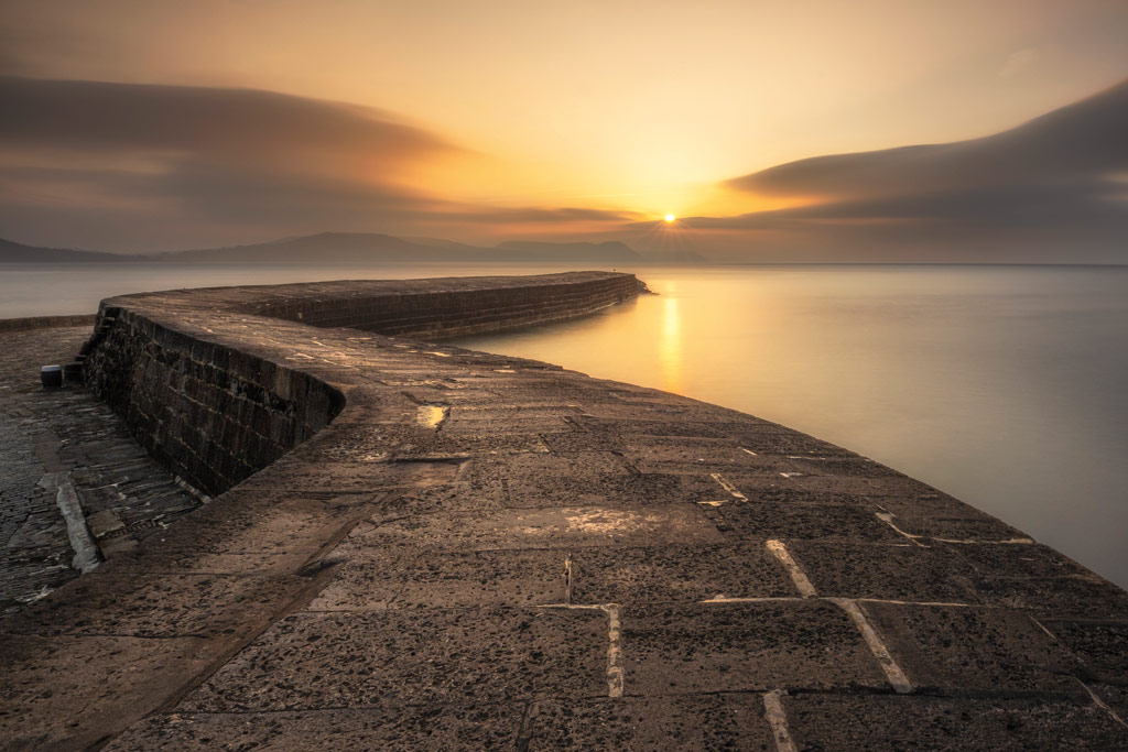 landscape with a winding stone pier leading out to the sea at sunset
