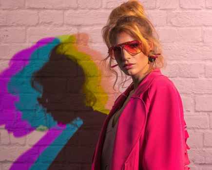 Woman in a pink jacket and shades standing by a pale pink brick wall with colour effect on her shawdow