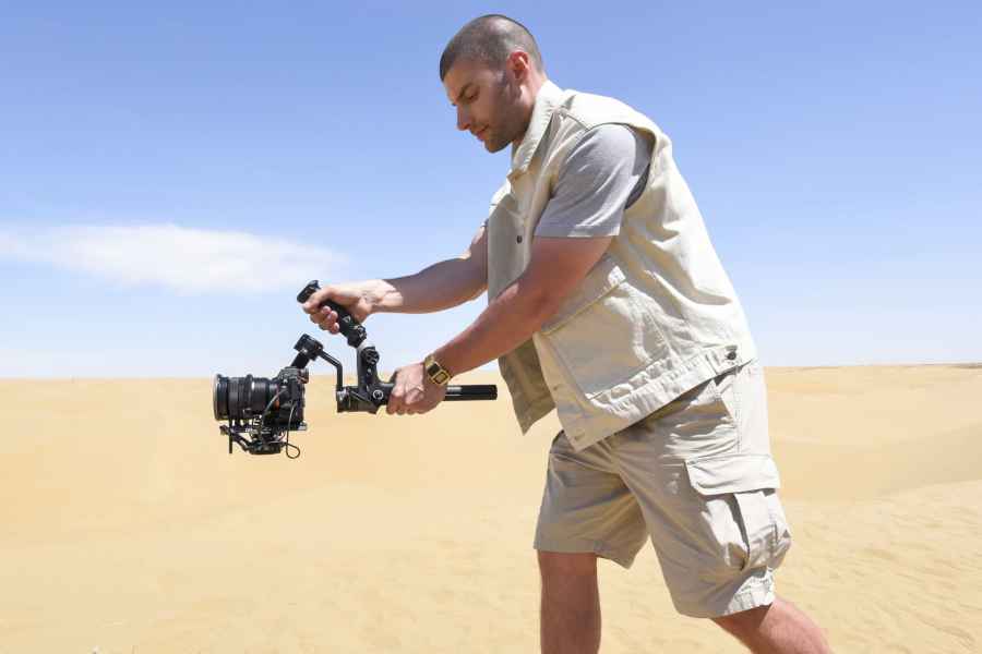 A man dressed in all beige filming with a camera in the desert