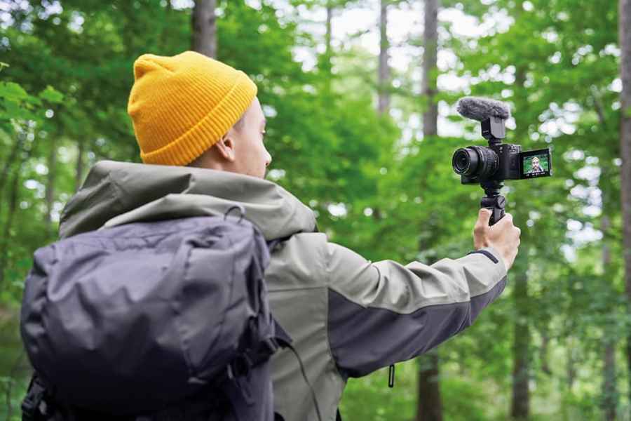 Man in a yellow knitted hat taking a selfie on a black camera in a green woodland area