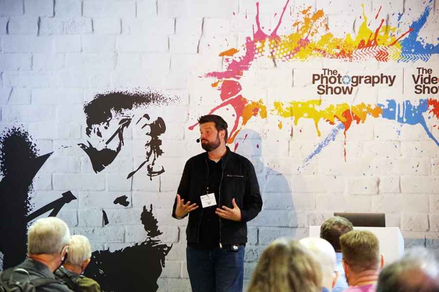 Man in black presenting to a crowd at the Photography show