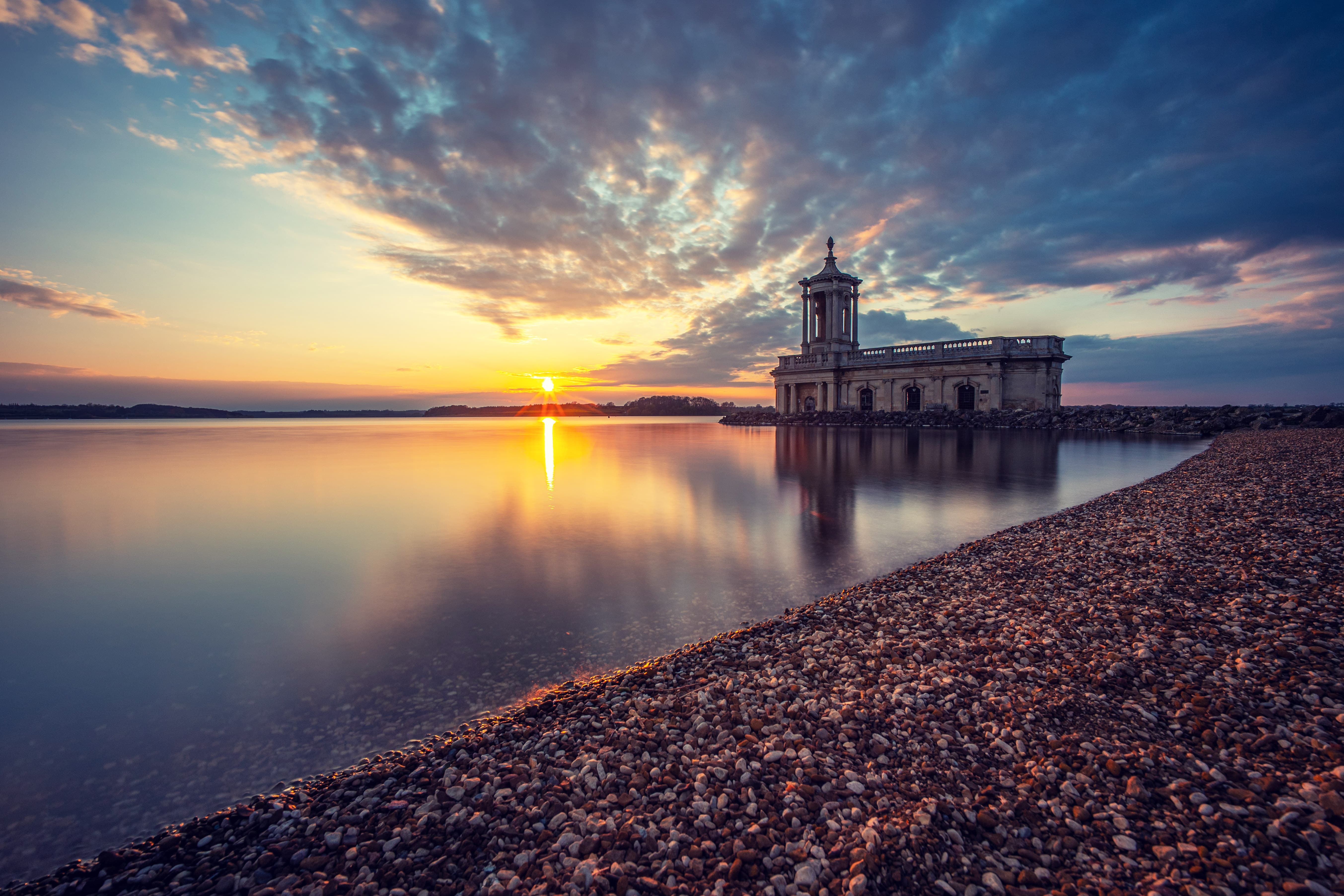 Colourful sunset with wispy clouds mirrored on a lake. A pebbly shore leads the eye to an interesting old building.