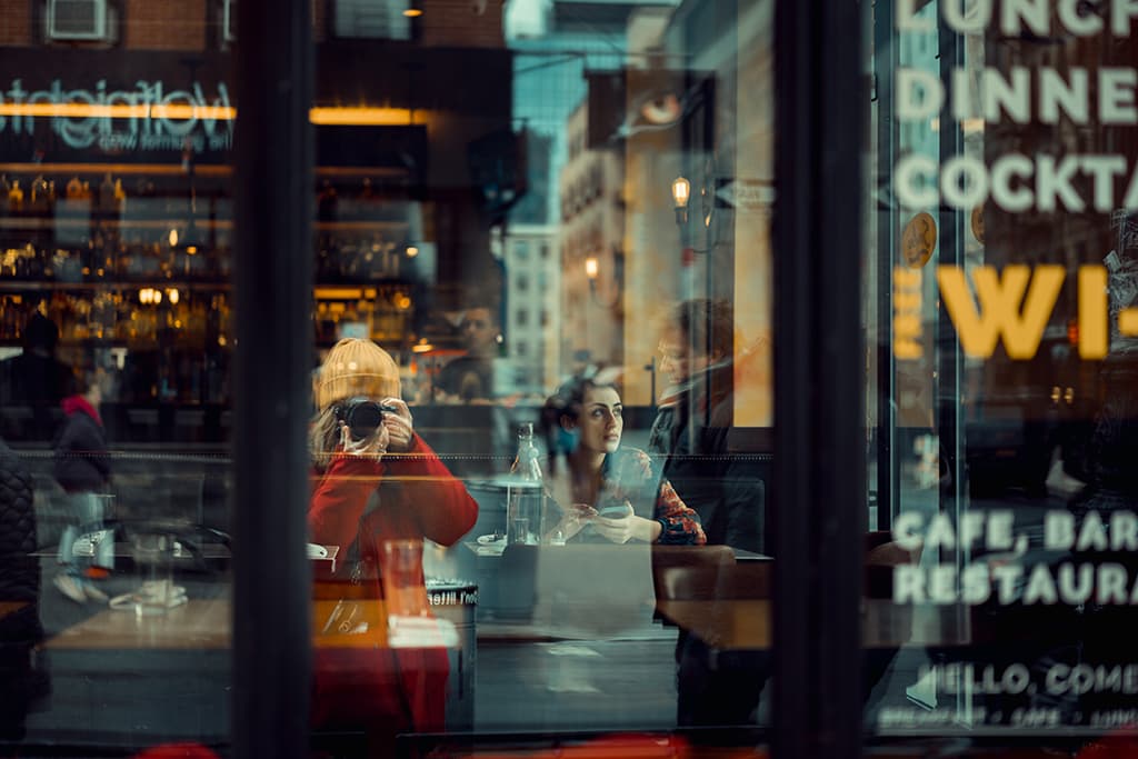 person in a yellow hat and red jumper photographs the reflection of the street on a shopwindow, inside the shop a woman sitting at a table 