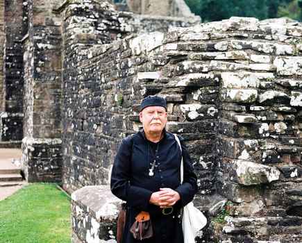 Man with a moustache in black top and hat stood infront of stone wall