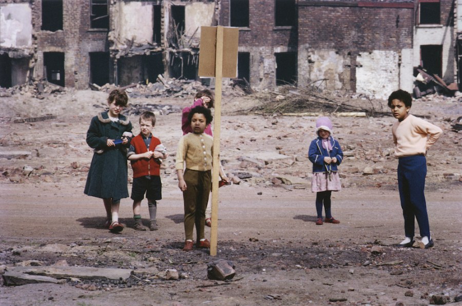 Street Scene, Hulme, Manchester. A group of children standing by a sign in front of a 'cleared' row of houses, the shells of houses due for demolition behind, 1965. Image: Shirley Baker Photography © Nan Levy for the Esta