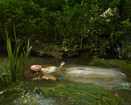 Girl in a white dress floating on back in forest water surrounded by reeds and flowers