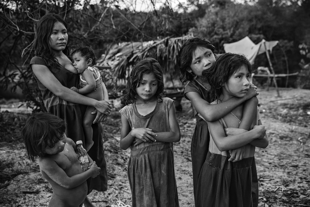 Women and children from the Pirahã community, standing next to their camp on the banks of the Maici River, watch drivers passing by on the Trans-Amazonian highway hoping to be given snacks or soft drinks, Humaitá, Amazon, Brazil, on 21 September 2016