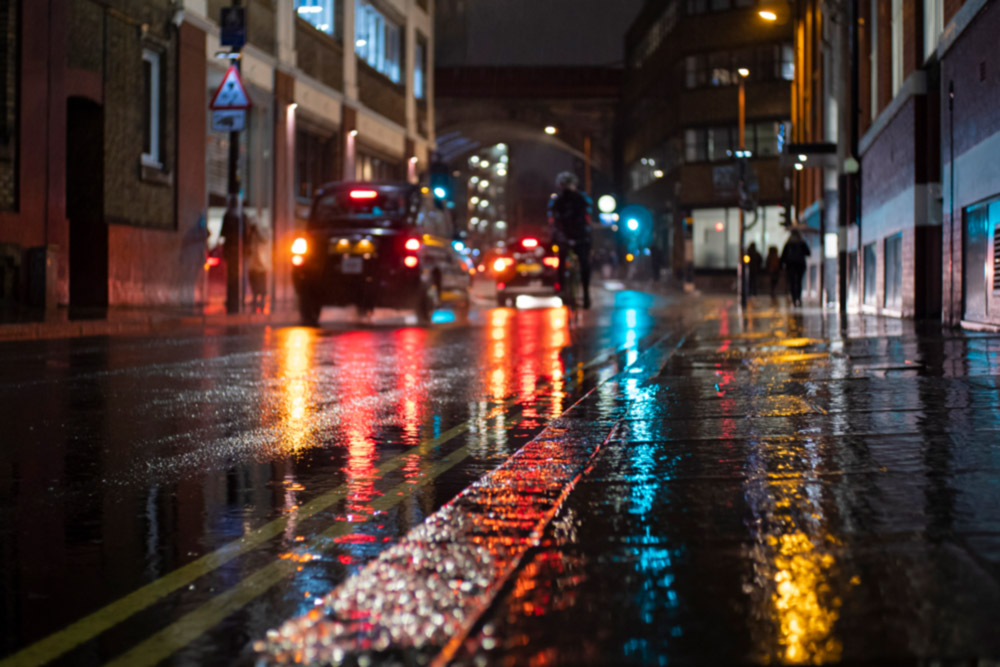 Cityscape, cars passing by on a rainy night, lights reflecting on the wet pavement, photographed from a very low angle.