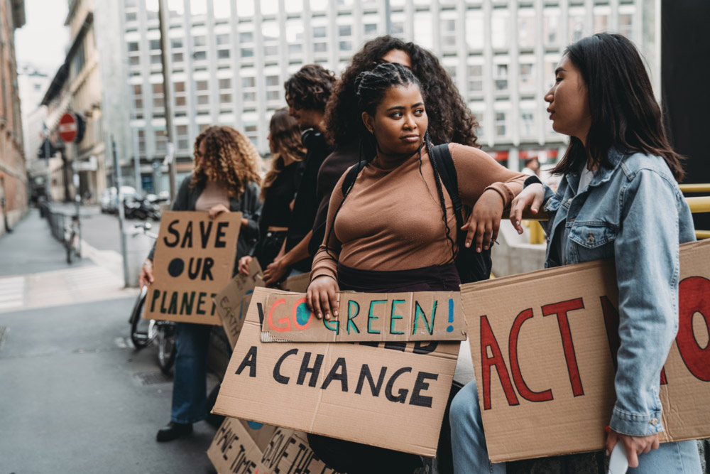 A group of young people lean against a railing on the street holding cardboard signs "Save our planet" "Go green" "`change" "act now"