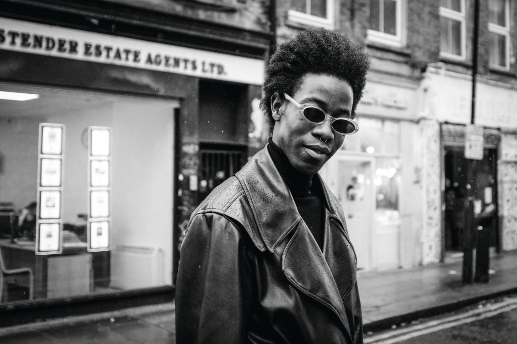 black and white image, a man in a leather jacket and black sunglasses looks at the camera