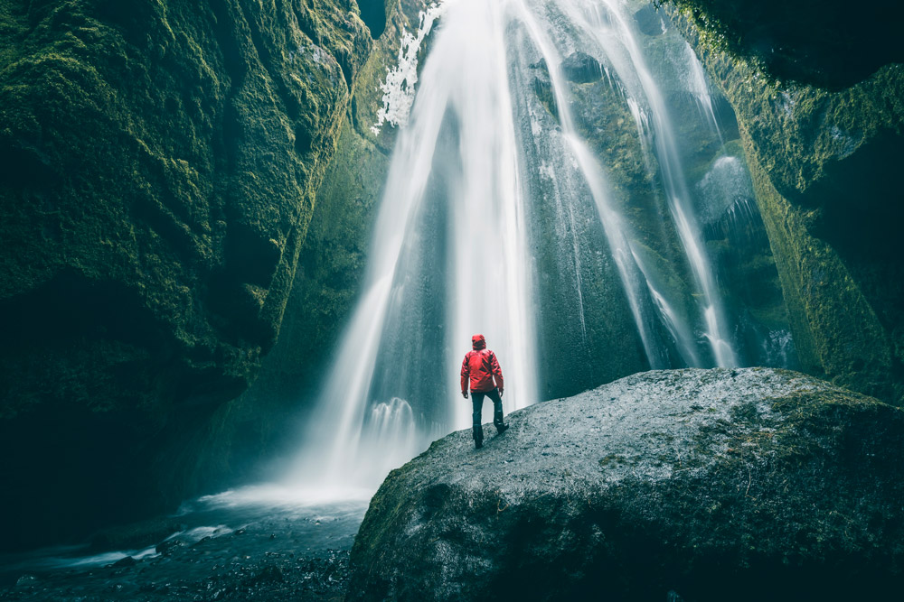 long exposure shot of a waterfall surrounded by mossy rock walls, a man in a red coat stands in the centre of the image dwarfed by the size of the waterfall and scenery. Credit: (C) Marco Bottigelli, Getty Images