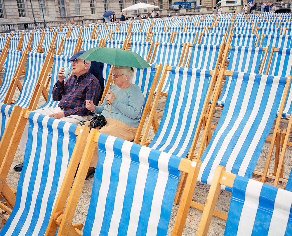 Elderly couple sat on blue and white striped beach chairs under green umbrella