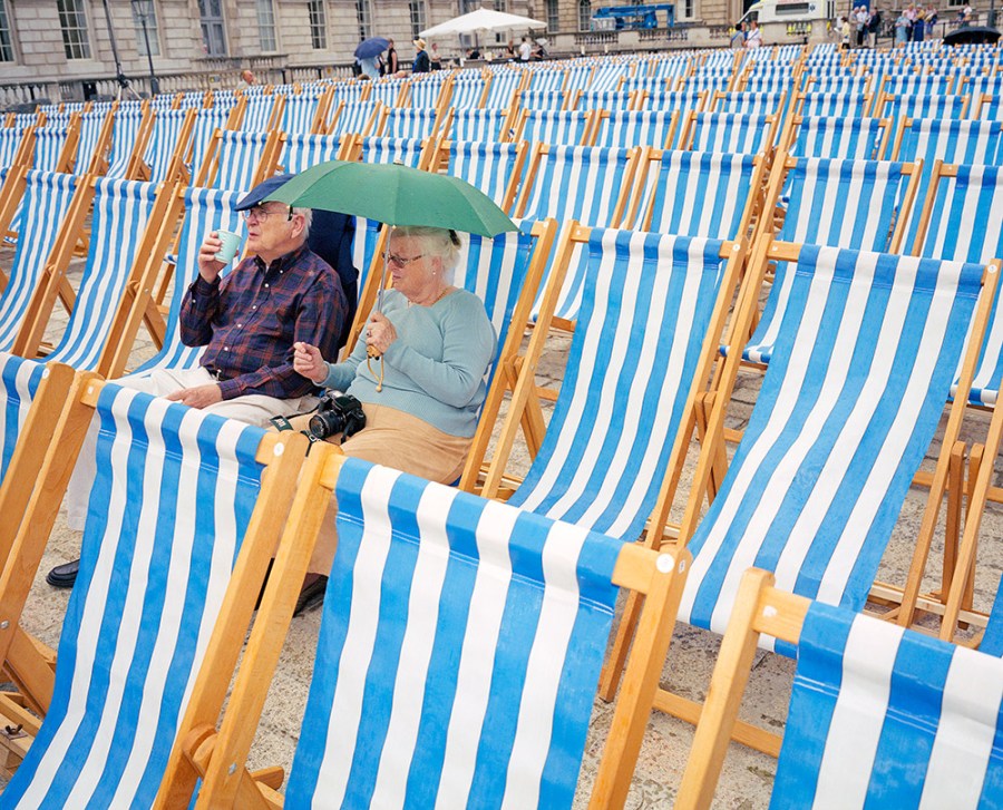 Elderly couple sat on blue and white striped beach chairs under green umbrella