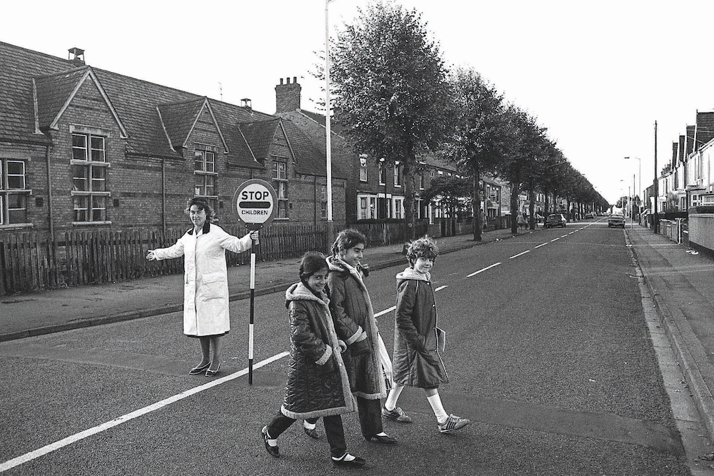 Lollipop lady Gloria Steele sees (from left to right) Mazia Ahmad, Gulfraz Umar (nee Malik) and Tanya Porter across the road at Queen's Drive School, 1984. © Chris Porsz