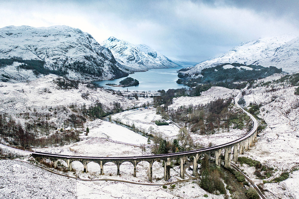 drone photograph overlooking snowy mountain scene with bridge and lake