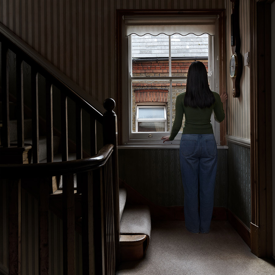 girl in long green top and blue jeans looks out of hallway window onto another house westminster