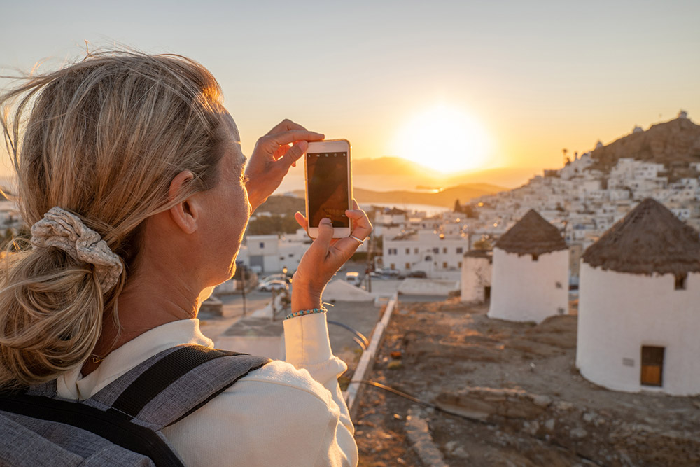 young woman travelling taking a photo of a sunset on greek island for social media