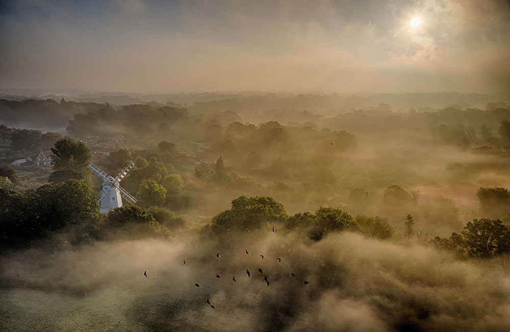 drone looking over countryside landscape with windmill and mist on the horizon