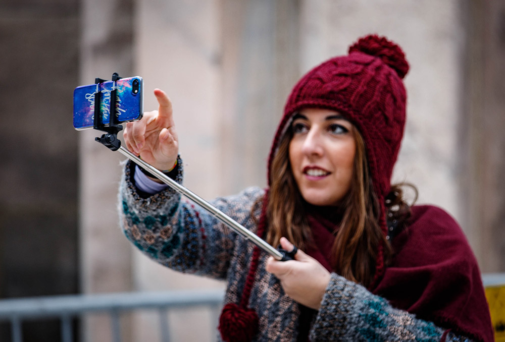 a woman in a red bobble hat takes a selfie using a smartphone on a selfie stick touching screen to manual focus image