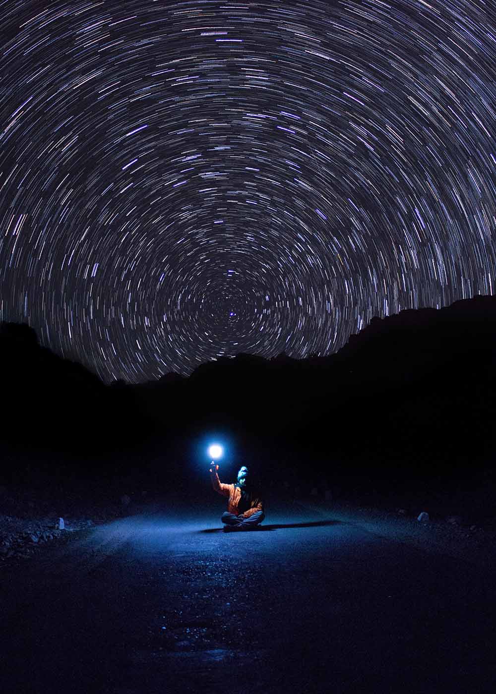 Night sky with circular star trails with silhouette of a hill. In the foreground a person sits on the road cross legged holding up a lamp.