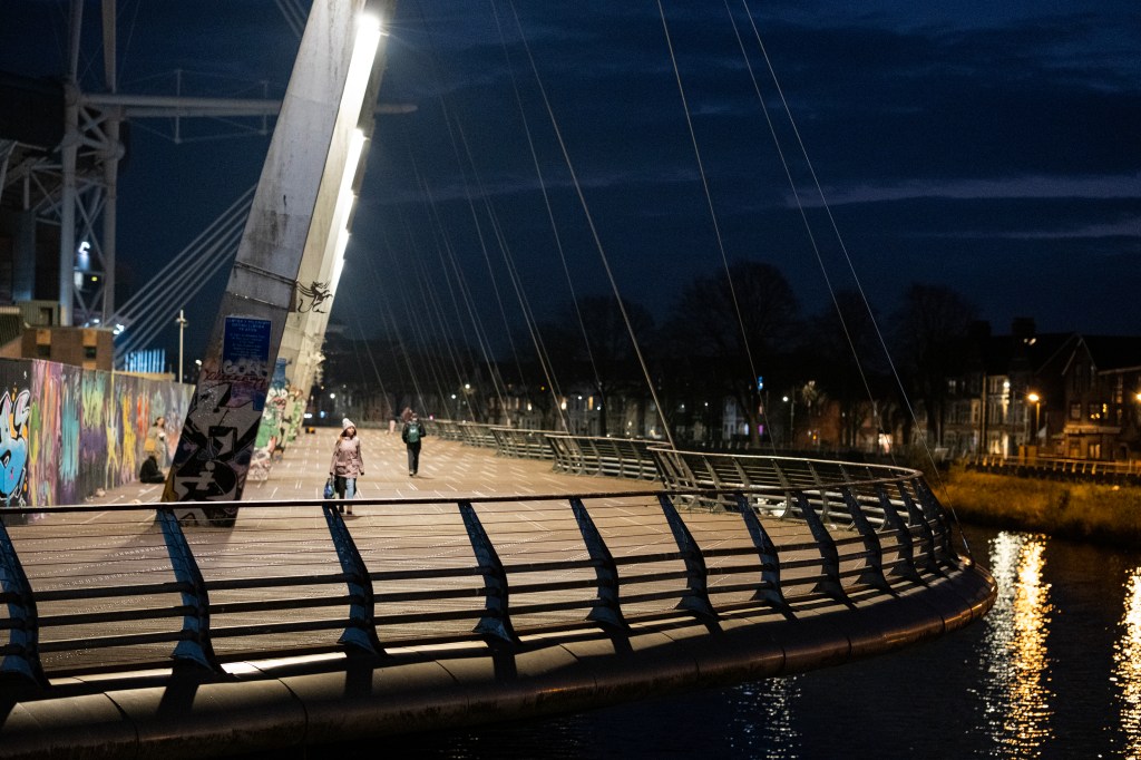 Two people walking on a wide bridge at night, on the left graffities on the wall, on the right houses on the bank of the river.