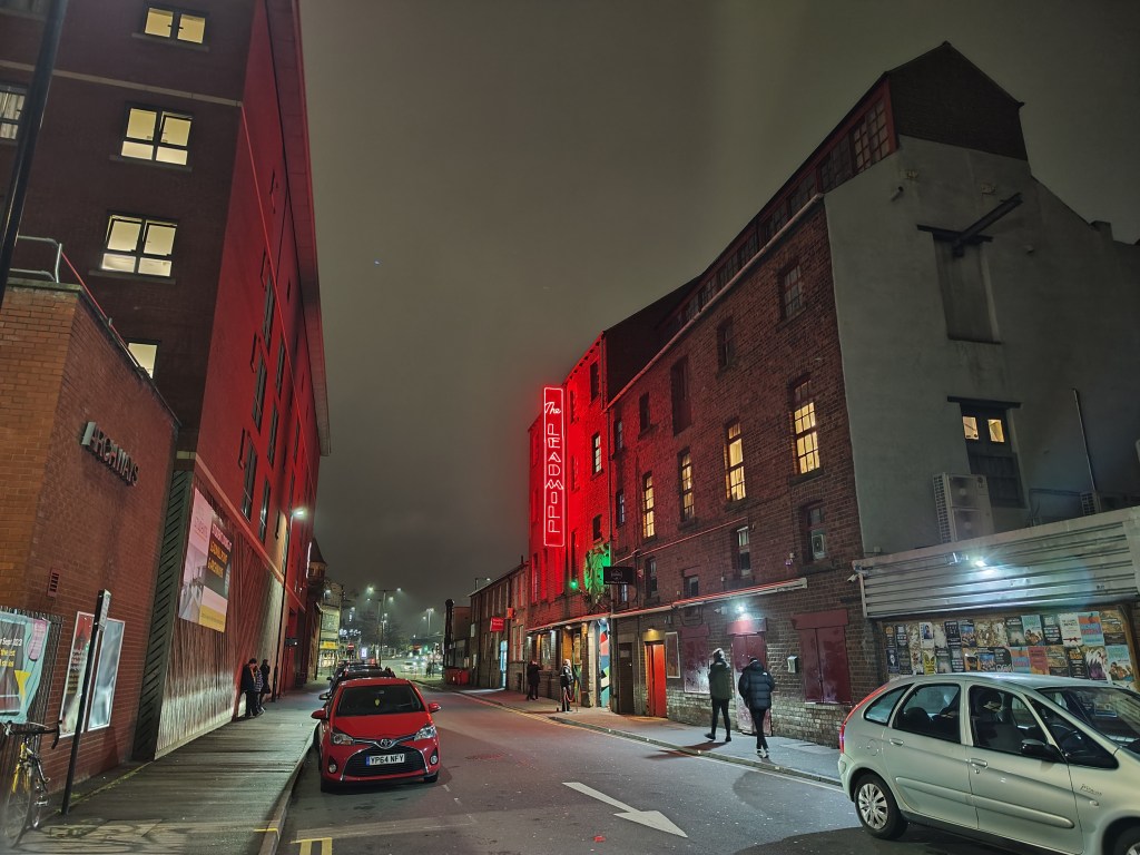 Taken with the main wide camera on the Xiaomi 12T Pro using the night mode. A street at night with three and four storey brick buildings, cars parking and a few people walking by, a big red neon sign on the wall says " the leadmill"