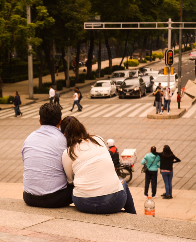 A couple sits on the steps of El Angel de la Independencia statue in Mexico City facing a busy street, candid portraits