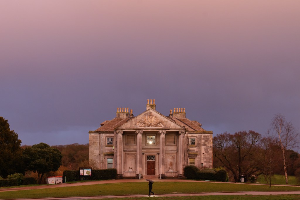 Panasonic Lumix S5II A mansion in a spring storm at sunset, a person in the middle of the frame a man walks by with an umbrella 