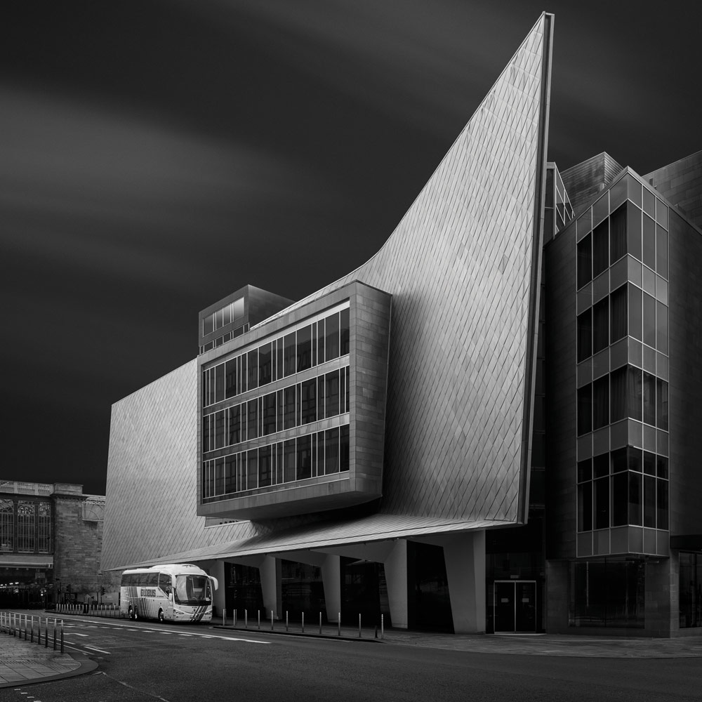 Black and white building photography, Billy Currie. Long exposure of a modern building with tiled facade, a white bus parked in front gives contrast to the image.