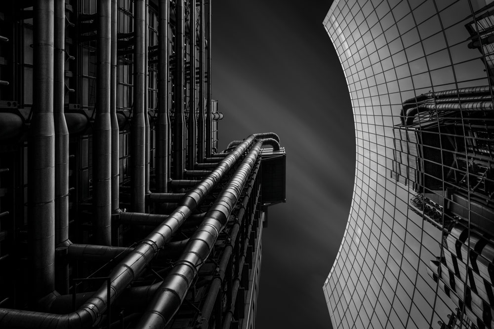 Black and white building photography, Billy Currie. Looking up a curved glass surfaced building that reflects the view of another building in front with dozens of pipes criss-crossing on its surface.