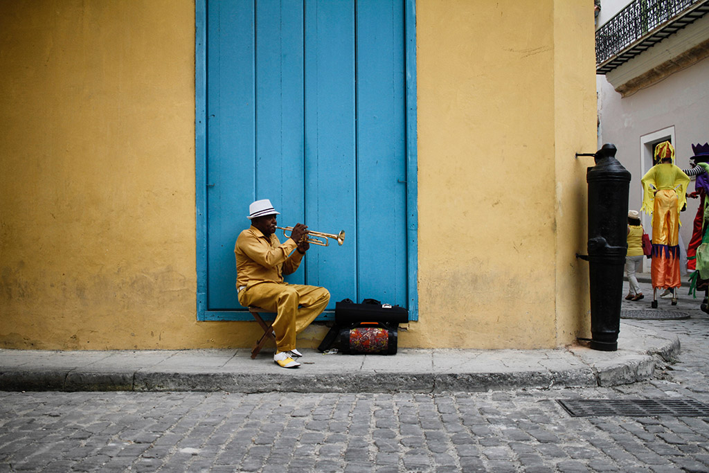 A man wearing yellow suit sat in front of a blue door on the street seen playing a trumpet in Havana