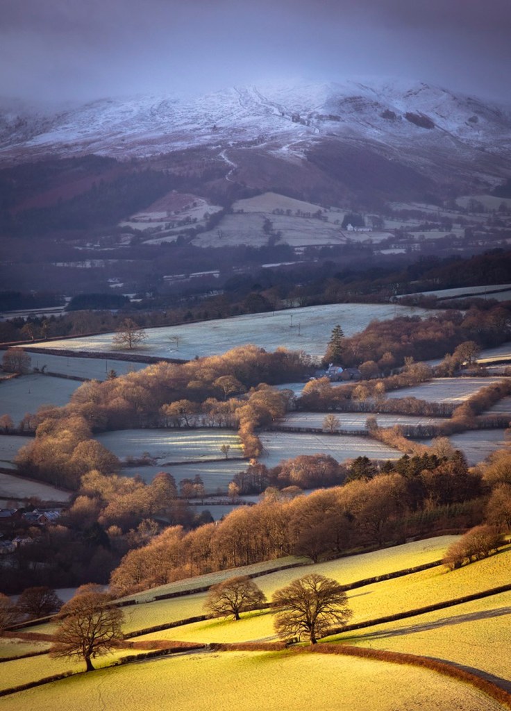 Landscape Photographer of the Year 2022 winner Will Davies image of Brecon Beacons National Park in Wales, from snowy, misty mountains far in the background the landscape transitions to frosty fields then to illuminated sunny land