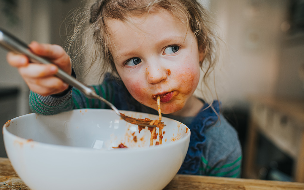 young girl eating spaghetti bolognese from a white bowl food around her mouth
