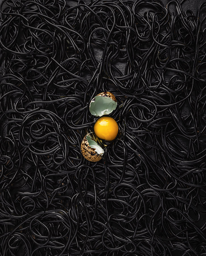 broken egg showing yellow yolk on top of black spaghetti Alone © Bobby Cortez | Pink Lady® Food Photographer of the Year 2023