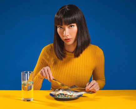dark haired woman in yellow jumper poses with knife and fork on yellow table against blue background