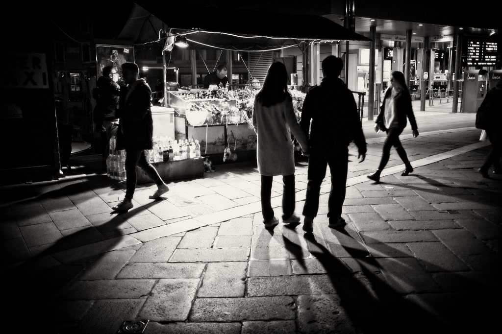 People walking by a street vendors stall at night. The stall's lights illuminate them and create silhouettes and long shadows. Black and white image.